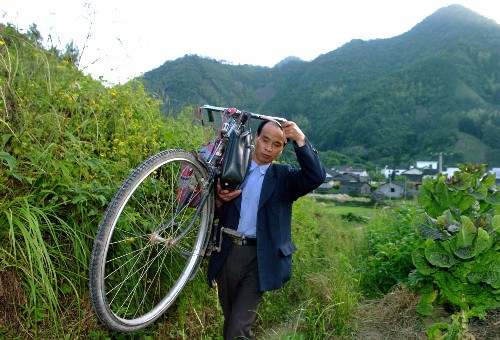 教师汪来九从自己住的村庄到十多里外的毛田小学去上课。由于交通不便，他还要扛着自行车走一段山路。